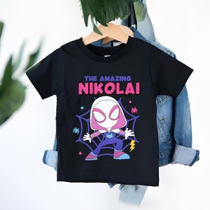 Puede incluir: Camiseta negra con una ilustraci&oacute;n de dibujos animados de un superh&eacute;roe y el texto "THE AMAZING NIKOLAI". El personaje est&aacute; en una telara&ntilde;a con ara&ntilde;as. La camiseta est&aacute; colgada en una percha de madera contra una chaqueta vaquera.