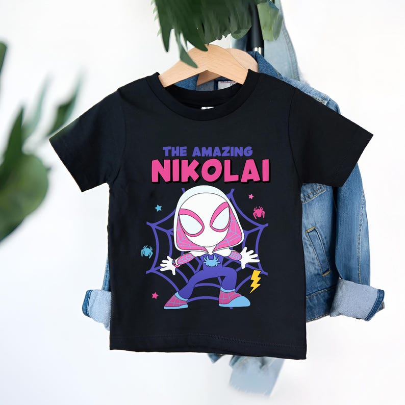 Puede incluir: Camiseta negra con una ilustraci&oacute;n de dibujos animados de un superh&eacute;roe y el texto "THE AMAZING NIKOLAI". El personaje est&aacute; en una telara&ntilde;a con ara&ntilde;as. La camiseta est&aacute; colgada en una percha de madera contra una chaqueta vaquera.