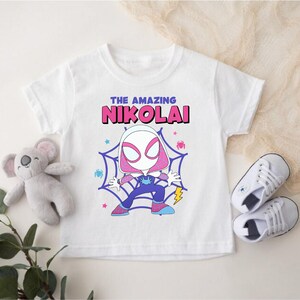 Puede incluir: Camiseta blanca con un gr&aacute;fico de dibujos animados de un superh&eacute;roe en un dise&ntilde;o de telara&ntilde;a. El texto "THE AMAZING NIKOLAI" est&aacute; impreso encima del personaje. La imagen incluye zapatillas blancas y un peluche de koala.