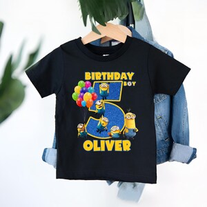May include: Black t-shirt featuring a colorful graphic of Minions and balloons. The text reads "BIRTHDAY BOY" and "OLIVER" with a large number "5". The design has yellow and blue accents, perfect for a child's birthday celebration.