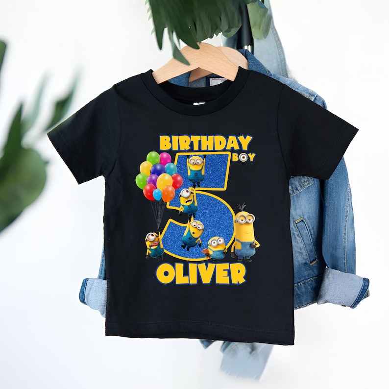 May include: Black t-shirt featuring a colorful graphic of Minions and balloons. The text reads "BIRTHDAY BOY" and "OLIVER" with a large number "5". The design has yellow and blue accents, perfect for a child's birthday celebration.