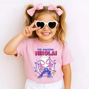 Puede incluir: Una camiseta rosa con el texto "THE AMAZING NIKOLAI" en morado y rosa. La camiseta presenta un dibujo animado de un personaje de superh&eacute;roe en una telara&ntilde;a. La ni&ntilde;a lleva gafas de sol en forma de coraz&oacute;n y lazos rosas.