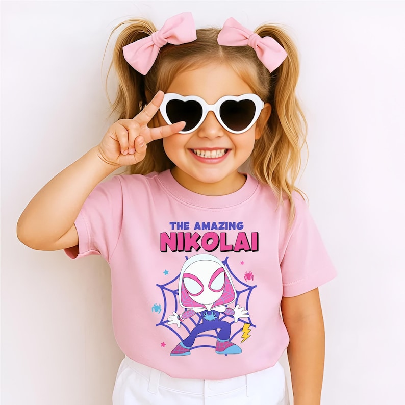 Puede incluir: Una camiseta rosa con el texto "THE AMAZING NIKOLAI" en morado y rosa. La camiseta presenta un dibujo animado de un personaje de superh&eacute;roe en una telara&ntilde;a. La ni&ntilde;a lleva gafas de sol en forma de coraz&oacute;n y lazos rosas.