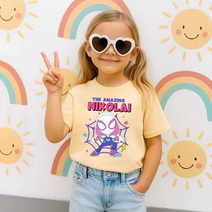 Puede incluir: Una camiseta amarilla clara con el texto "THE AMAZING NIKOLAI" sobre un dibujo animado de un superh&eacute;roe. La ni&ntilde;a lleva gafas de sol en forma de coraz&oacute;n y vaqueros azules. El fondo presenta arco&iacute;ris y soles sonrientes.
