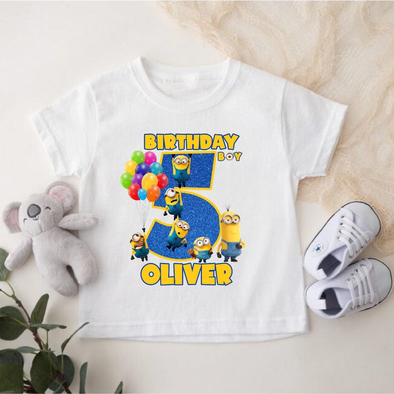 May include: White t-shirt featuring a colorful design for a child's birthday. The design includes the text "BIRTHDAY BOY" and "OLIVER", along with the number "5" and several Minion characters. A koala plush and white sneakers are also visible.