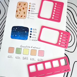 Può includere: Un foglio di carta bianco con campioni di colore, un'illustrazione di notte stellata e il testo "Sfumature d'autunno". Tre guide per palette di colori in plastica rosa sono sulla carta. Vari pennarelli colorati sono sullo sfondo.