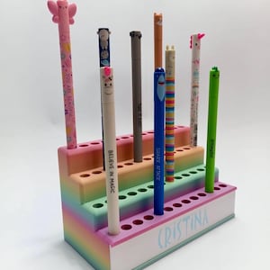 May include: A multi-tiered pen holder in pastel colors, holding several pens with animal-themed toppers. The holder has a rainbow gradient and the name "CRISTINA" is written on the front. The pens have various designs and the text "YOU ARE BEAUTIFUL" and "BELIEVE IN MAGIC".