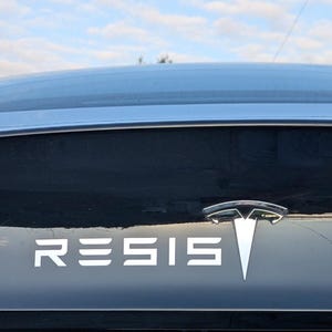 Op de afbeelding: De achterkant van een zwarte Tesla met een wit Tesla-logo en het woord "RESIS" in witte letters.