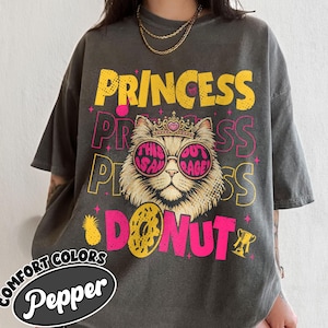May include: Dark gray t-shirt with a graphic of a cat wearing sunglasses and a crown, with the words "PRINCESS DONUT" in yellow and pink. The shirt also features a pineapple and a trophy.