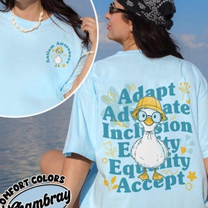 May include: Light blue t-shirt with a graphic of a duck wearing glasses and a yellow hat. The back of the shirt has the words "Adapt, Advocate, Inclusion, Equity, Accept". The front of the shirt has a small graphic with the words "Autism Awareness".