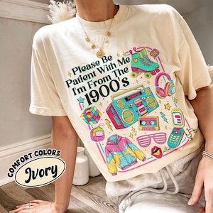May include: Ivory-colored t-shirt with a colorful 1990s graphic. The graphic includes a boombox, roller skates, a Rubik's cube, and the text "Please Be Patient With Me I'm From The 1900's". The shirt also has the text "Comfort Colors Ivory".