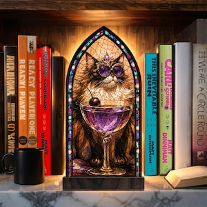 May include: A gothic-arched stained glass lamp featuring a fluffy cat wearing purple sunglasses and a crown, sitting behind a martini glass with a cherry. The lamp has a dark wooden base and emits a warm glow, positioned between several books.