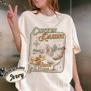 Può includere: T-shirt color avorio con grafica "Chicken Raising & Jesus Praising". Il design presenta un pollo, balle di fieno, una croce e una piccola capanna. La maglietta fa parte della collezione Comfort Colors.