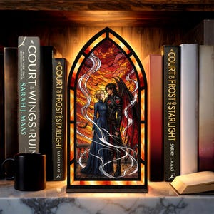 May include: An arched, illuminated display featuring a stained-glass style illustration of a couple standing on a balcony against a fiery orange sky. The display is flanked by books titled A Court of Wings and Ruin and A Court of Frost and Starlight.