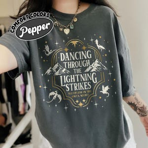 Pode incluir: Camiseta cinza escuro com o texto "Dancing Through The Lightning Strikes" em um design dourado e branco. A camisa apresenta ilustrações de montanhas e dragões. A marca "Pepper" está em um oval branco.