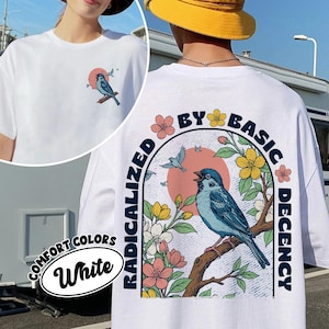 以下が含まれることがあります： 前面と背面に鳥のグラフィックが描かれた白いTシャツ。前面には、夕焼けを背景に枝にとまる青い鳥が描かれています。背面には、より大きな鳥のデザインと「RADICALIZED BY BASIC DECENCY」の文字があります。