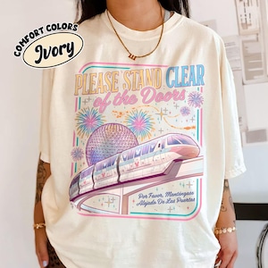 Op de afbeelding: Ivoor kleurig T-shirt met een kleurrijke afbeelding. De afbeelding toont een monorail, vuurwerk en de tekst "PLEASE STAND CLEAR of the Doors" in retro stijl. Onder de tekst staat de zin "Por Favor, Manténgase Alejado De Las Puertas".