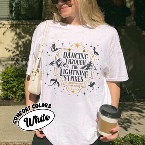 May include: White t-shirt with a graphic design featuring the text "Dancing Through The Lightning Strikes" and a mountain scene. The shirt also has the words "Sleepless in the Onyx Night". The shirt is labeled "Comfort Colors White".