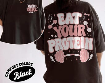 May include: Black t-shirt with the text "GYM GIRL" on the front and "EAT YOUR PROTEIN" on the back. The back design includes pink dumbbells and lightning bolts. The shirt is part of the Comfort Colors collection.