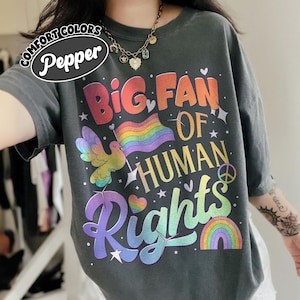 Puede incluir: Camiseta gris oscuro con el texto "Big Fan of Human Rights" en letras coloridas con temática de arcoíris. El diseño incluye una bandera arcoíris, una paloma sosteniendo una bandera, corazones, estrellas y un símbolo de la paz. La camiseta tiene un corte holgado.