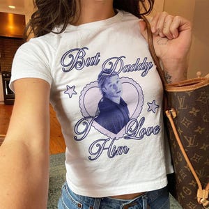 May include: A white short-sleeved t-shirt featuring a central graphic of a person inside a light purple heart frame. The text "But Daddy I Love Him" is printed in dark purple script around the heart, accompanied by two small star icons.
