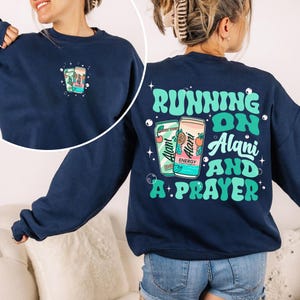 May include: Navy blue sweatshirt with a graphic of two energy drink cans and the text "Running on Alani and a Prayer" in teal and white. The front of the sweatshirt has a smaller version of the can graphic.