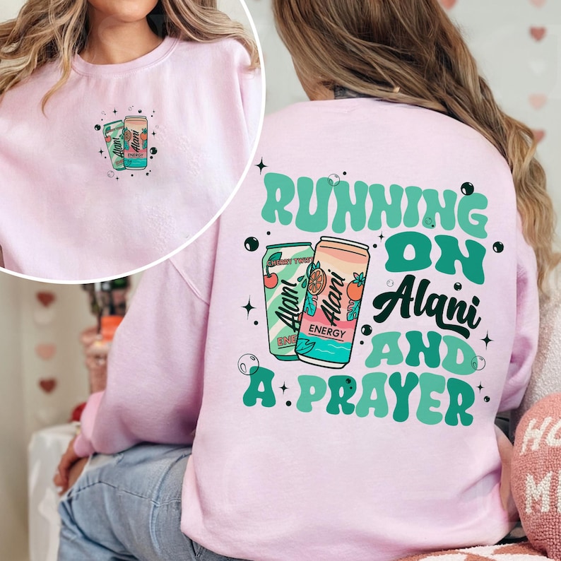 May include: A light pink sweatshirt with a graphic of two energy drink cans on the front and back. The back of the sweatshirt reads "RUNNING ON Alani AND A PRAYER" in green and white text. The front has a smaller version of the energy drink cans.