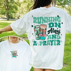 May include: White t-shirt with a graphic design featuring two beverage cans and the text "RUNNING ON Alani AND A PRAYER" in teal and black. The shirt also has a smaller graphic on the front.