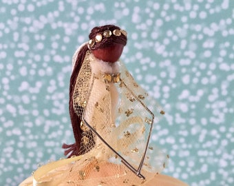 May include: A doll wearing a yellow dress with gold glitter and a brown hairpiece with rhinestones. The doll is wearing a sheer veil and has a brown face.