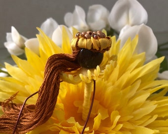 May include: A small brown and yellow doll with a brown tassel hairpiece sits on a yellow flower.