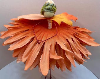 May include: A whimsical, handmade fairy figurine. The fairy has a small, round head with a green and yellow woven cap. The body is a large, orange flower with detailed petals. A red wire forms the arms and a small stone base.