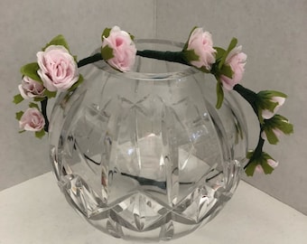 May include: A pink flower crown with green leaves. The crown is made of artificial flowers and is sitting on a clear glass vase.