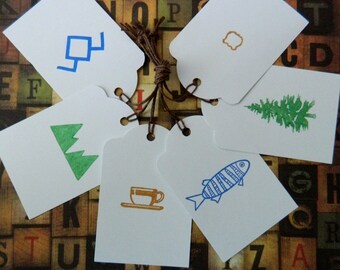 May include: Six white gift tags with different blue, green, and brown illustrations. The illustrations include a tree, a fish, a cup, a geometric shape, and a bell. The tags are tied together with twine.