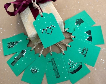 May include: Nine green gift tags with white snowflakes and various illustrations, including a mountain range, a tree, a cup of coffee, a pie, a log, a watering can, and a mountain range. The tags are tied together with twine.