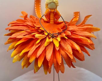 May include: A whimsical handmade fairy figurine. The fairy has a yellow yarn hair, an orange dress, and a skirt made of orange and yellow petals. The fairy is standing on a small gray stone base with wire legs. A decorative orange ribbon is attached.