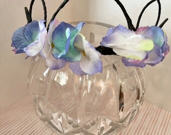 May include: A floral headband with blue, white, and purple flowers on a black wire frame. The headband is sitting on a clear glass vase.