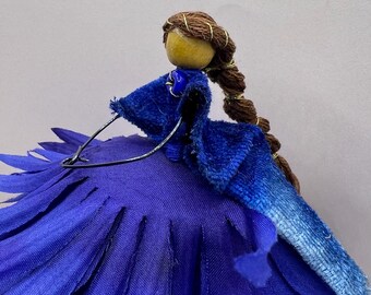 May include: A handcrafted fairy doll with a wooden head and brown braided hair adorned with gold thread. The doll wears a blue velvet dress and a large, layered blue fabric skirt. A wire frame supports the skirt.