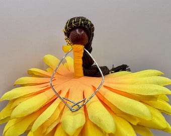 May include: A decorative figurine featuring a dark brown wooden bead head with braided dark brown yarn hair accented by gold thread. The body is wrapped in bright yellow yarn, and the skirt is made of layered yellow fabric petals.
