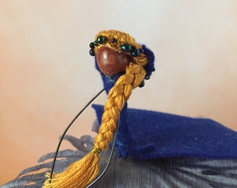 May include: A handmade doll with a blue felt cape, a braided yellow hair, and a crown of beads. The doll is sitting on a gray fabric base.