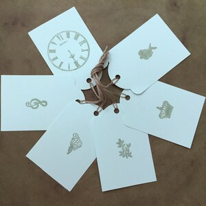 Puede incluir: Seis etiquetas de regalo blancas con diferentes diseños, que incluyen un reloj, una nota musical, una corona, una sirena, un corazón y una hoja. Las etiquetas están atadas con un cordel.