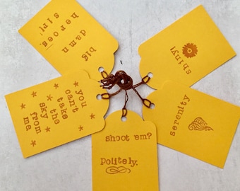 May include: Five yellow gift tags with brown string. The tags have different sayings on them, including "You can't take the sky from me", "shoot em? Politely", "serenity", "shiny", and "big damn heroes sir!"