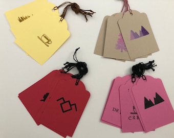 May include: A set of six gift tags with different designs. The tags are made of brown, yellow, pink, and red paper. The designs include a coffee cup, a mountain range, a tree, and a geometric shape. The tags are tied with twine.