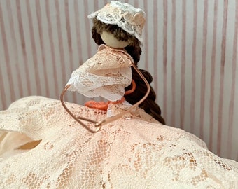 May include: A handmade doll with brown hair, wearing a white lace dress and a white lace hat. The doll is standing on a pink and white striped background.