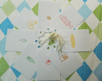May include: Eight white gift tags with various hand-drawn illustrations of food items, including a fish, corn, a pea pod, a tomato, bread, a pepper, a chicken, and an egg. The tags are tied together with a white string.