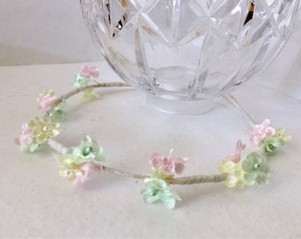 May include: A delicate floral headband with pastel pink, green, and yellow flowers. The headband is made of a thin, flexible wire and is adorned with small, pearl-like beads.