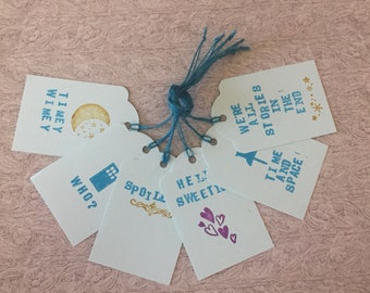 May include: A set of six white gift tags with blue and gold lettering. The tags feature quotes from the popular science fiction television series Doctor Who. The quotes include "Timey Wimey", "We're all stories in the end", "Hello Sweetheart", "Time and Space", "Who?" and "Spoil Me".