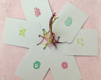 May include: A collection of light blue gift tags with various colorful stamped designs, including flowers, a paisley, and musical notes. The tags are tied together with a multicolored string, creating a visually appealing arrangement.
