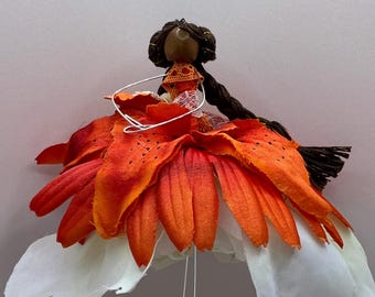 May include: A handcrafted doll featuring a dark brown yarn head and long braided hair. The doll wears a skirt made of layered orange and red tiger lily petals over white fabric petals, with a small orange lace bodice and thin wire arms.
