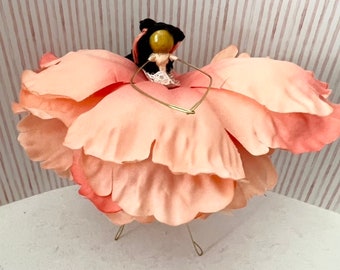 May include: A pink fabric flower with a doll in the center. The doll has a black head and a pink dress. The flower is on a wire stand.
