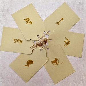 May include: Six beige gift tags with gold foil designs. The designs include a key, a fox, a swirl, a mermaid, a leaf, and a teapot.